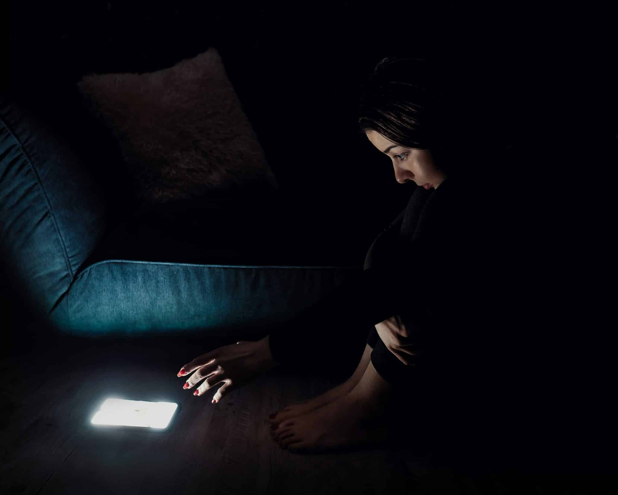 A woman sitting on the floor in a dark room, staring at her phone with eyes wide open and her hand reaching towards the bright screen. The image captures the mental strain and effects of doomscrolling on mental health.