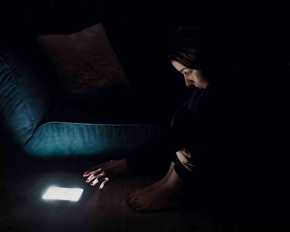 A woman sitting on the floor in a dark room, staring at her phone with eyes wide open and her hand reaching towards the bright screen. The image captures the mental strain and effects of doomscrolling on mental health.