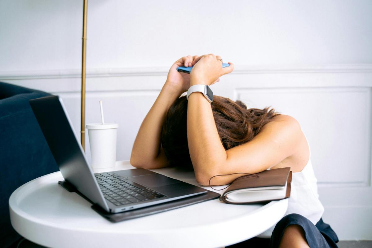 Woman in a white top resting her head on a table while holding a smartphone, appearing emotionally exhausted or digitally overwhelmed.