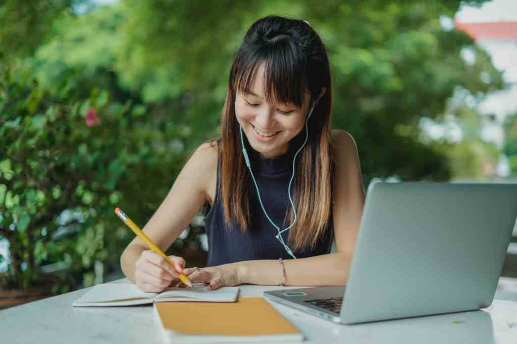 Young Asian woman writing in her notebook with laptop and headphones outdoors