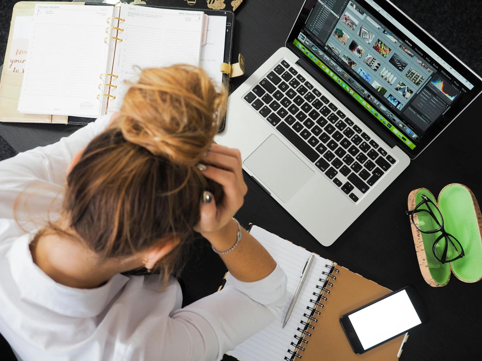 A frustrated woman sits at her desk with hands on her head, overwhelmed by work, surrounded by a laptop and documents, reflecting stress, burnout, and nervous system overload.